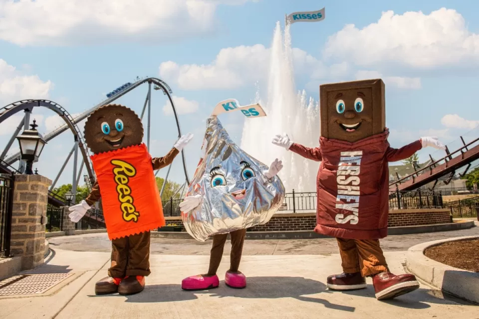 Three people are dressed in Reese's, Hershey's Kisses, and Hershey's Milk Chocolate Bar costumes in an amusement park with a roller coaster and fountain in the background